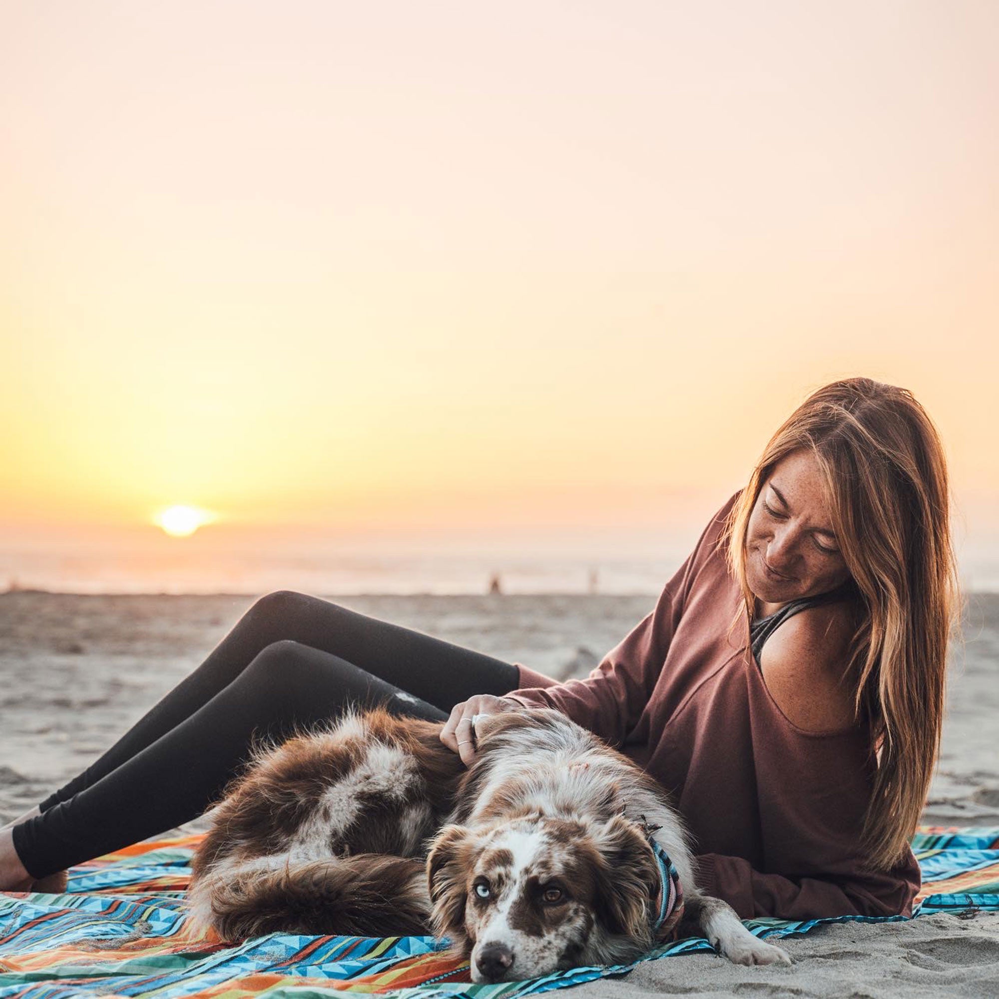 woman sits on adventure sheet on the beach with dog and klean kanteen waterbottle sunset showing a little shoulder