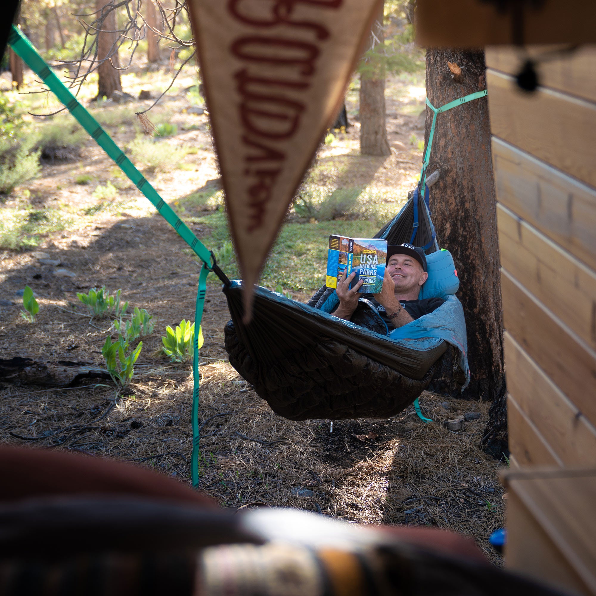 Man reading in Evolution hammock with a smile on his face outside van life hanging on tree trunk with pendant and forest