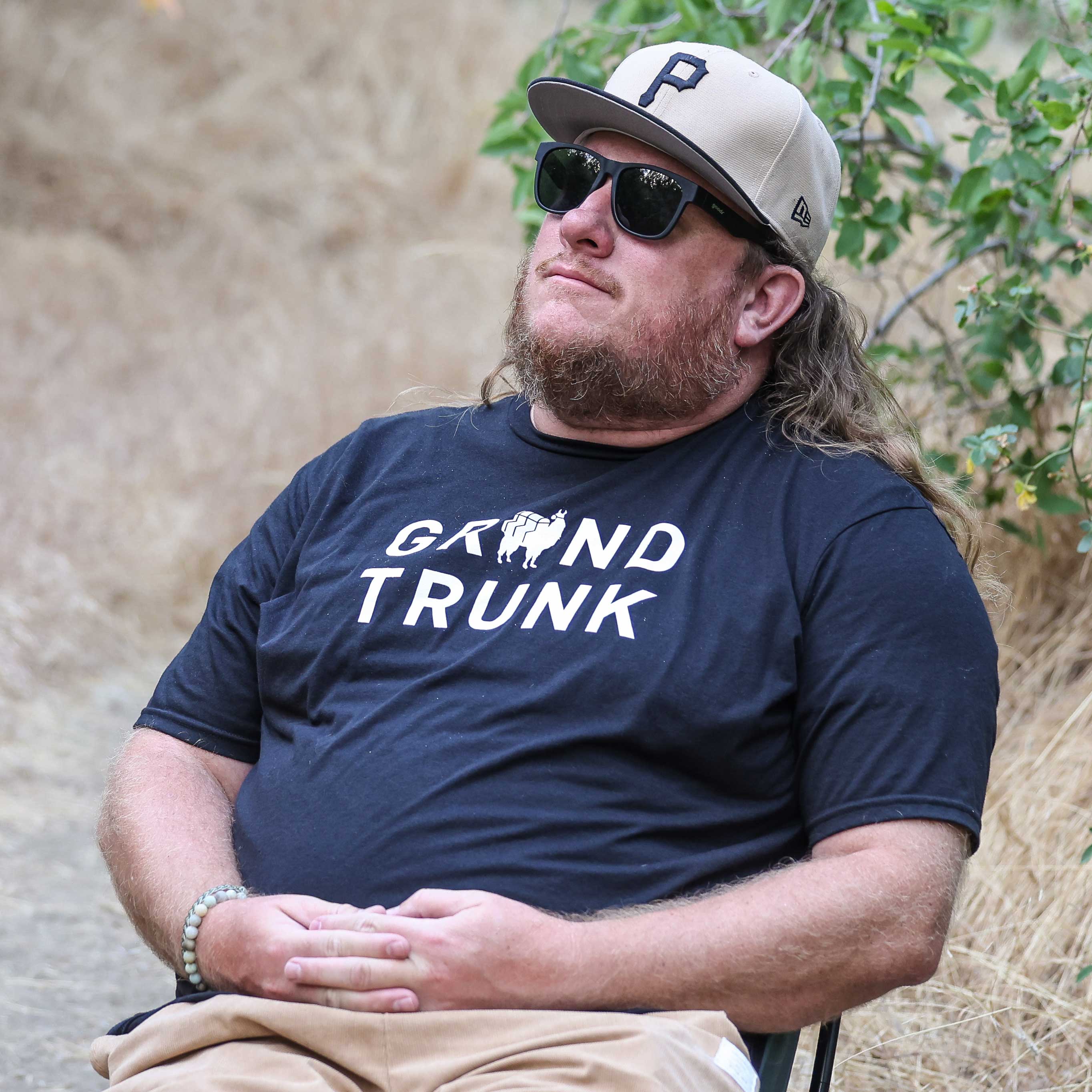 Man wearing a navy 'Grand Trunk' t-shirt with a cap and sunglasses, sitting outdoors.