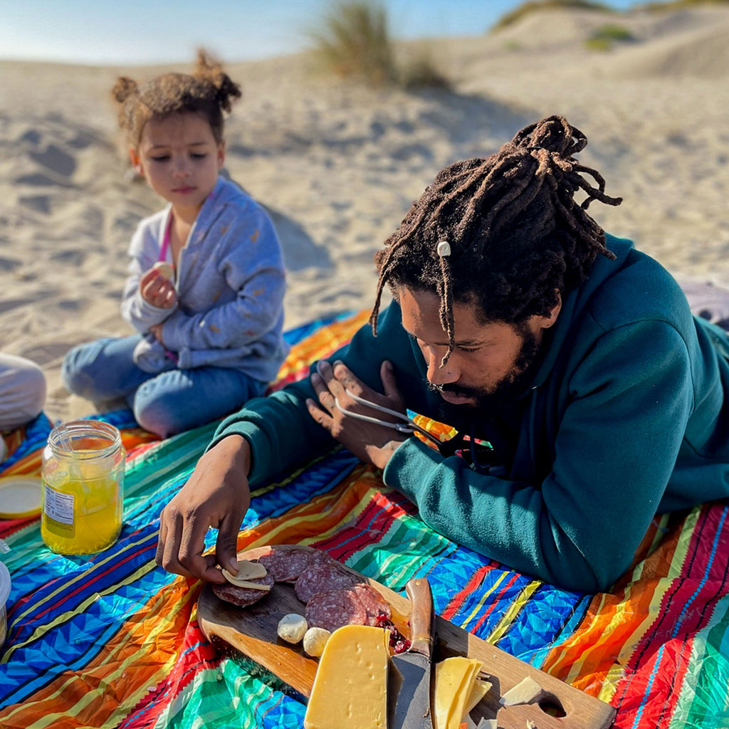 man eats from charcuterie board on the beach with the adventure sheet and his family children child