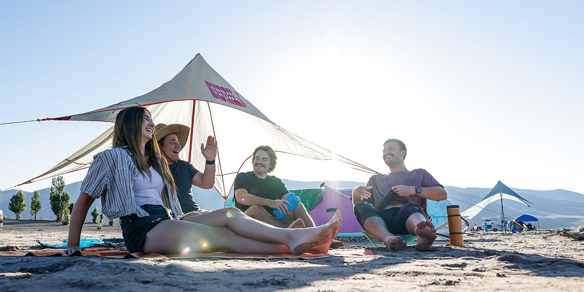 Group of homies hangs out on the beach underneath the ShadeCaster 2 with Grand Trunk chairs and parasheet and microfiber high tide low tide towels paddle boards SUP