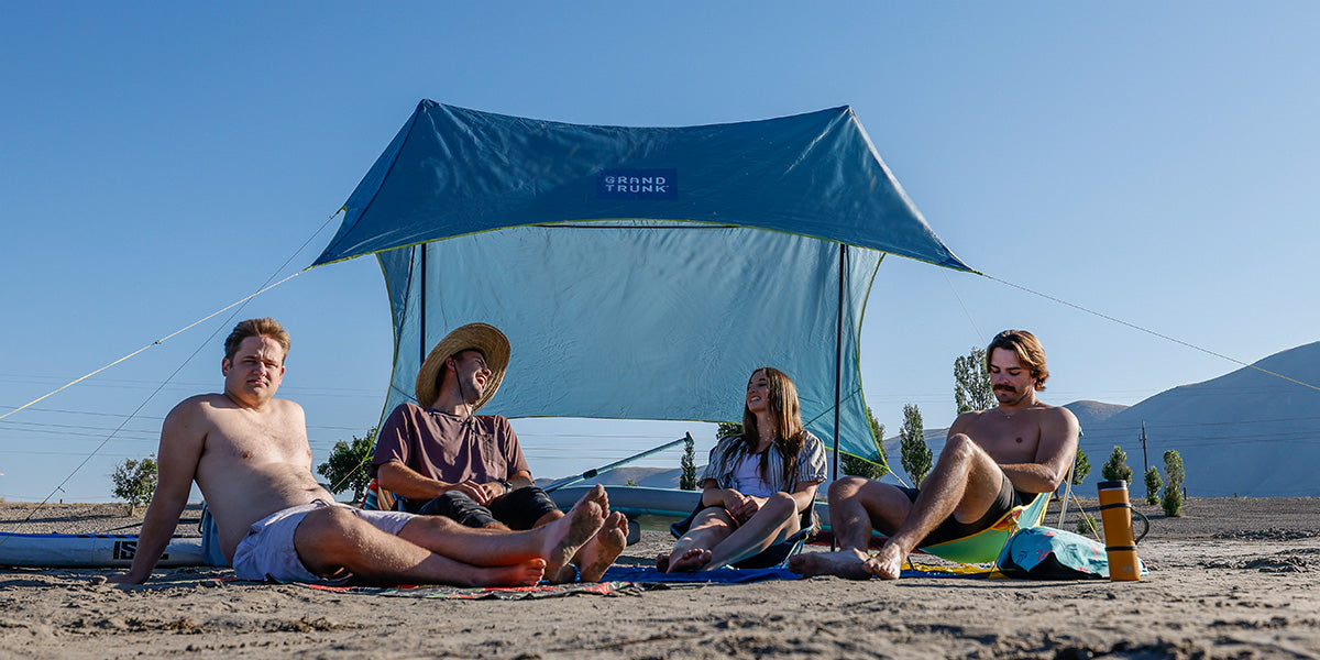 Group of homies hangs out on the beach underneath the ShadeCaster 4 with Grand Trunk chairs and parasheet and microfiber high tide low tide towels paddle boards SUP