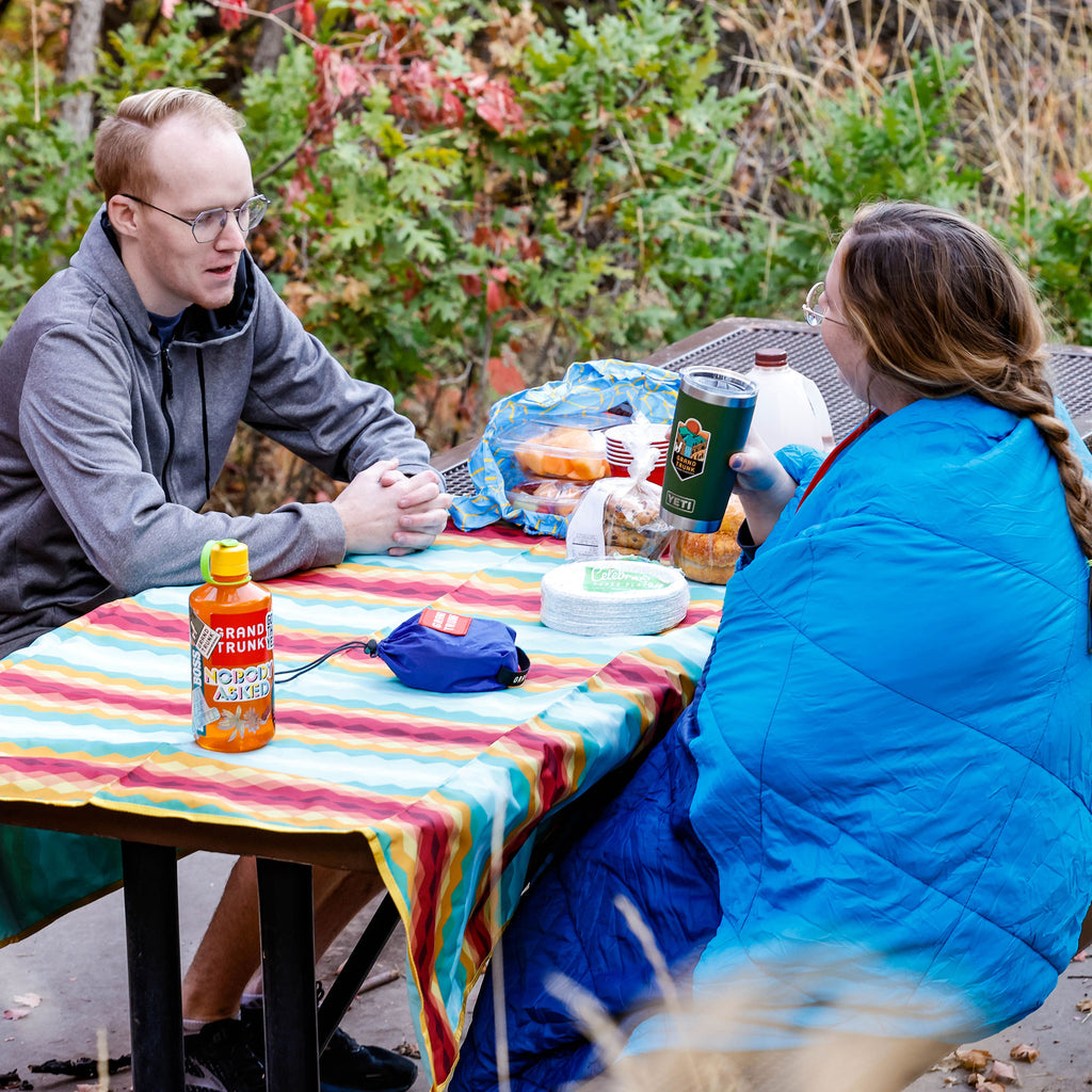 Couple using Grand Trunk Meadow Mat as table cloth on picnic table and bundling up in the thermaquilt