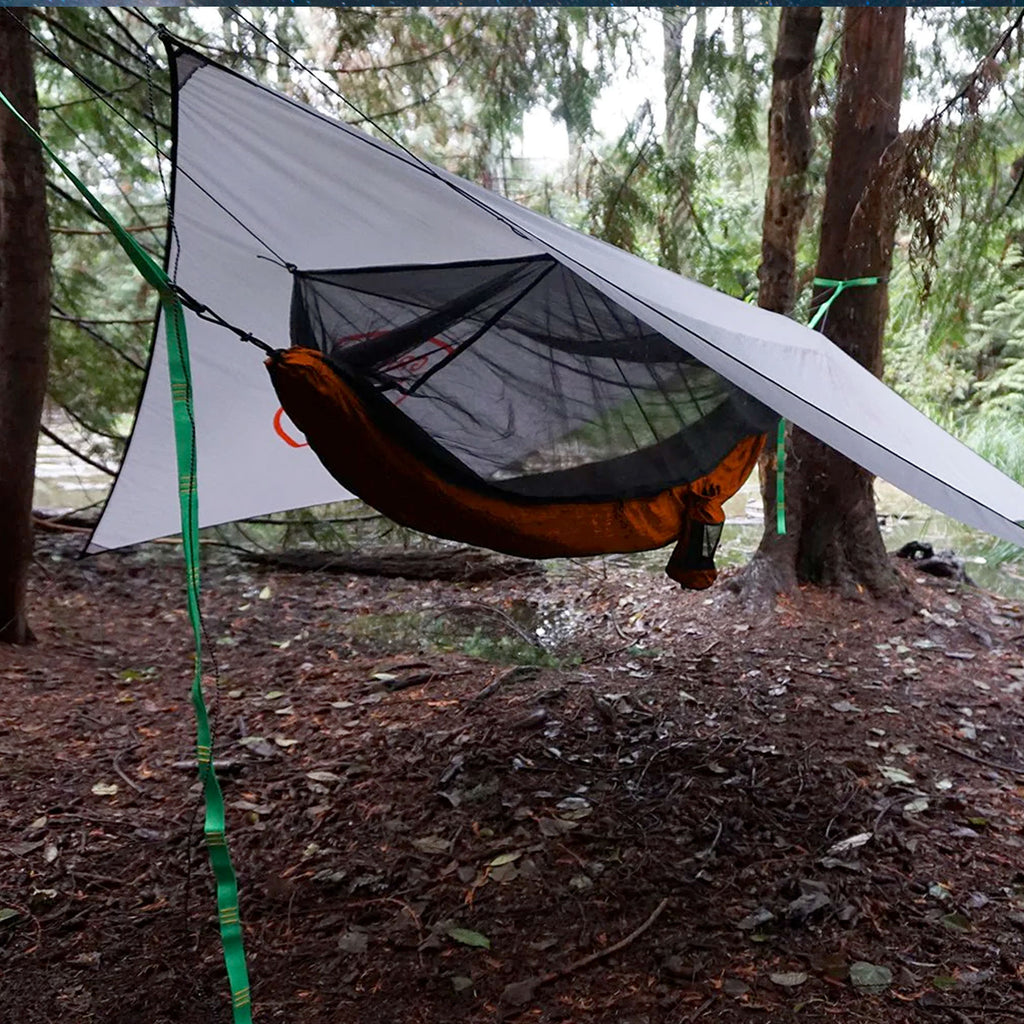 the abrigo all weather shelter setup with tarp and mosquito free hammock held up with trunk straps in the forest outdoor adventure
