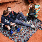 Three children sitting on a patterned blanket in a natural rock setting
