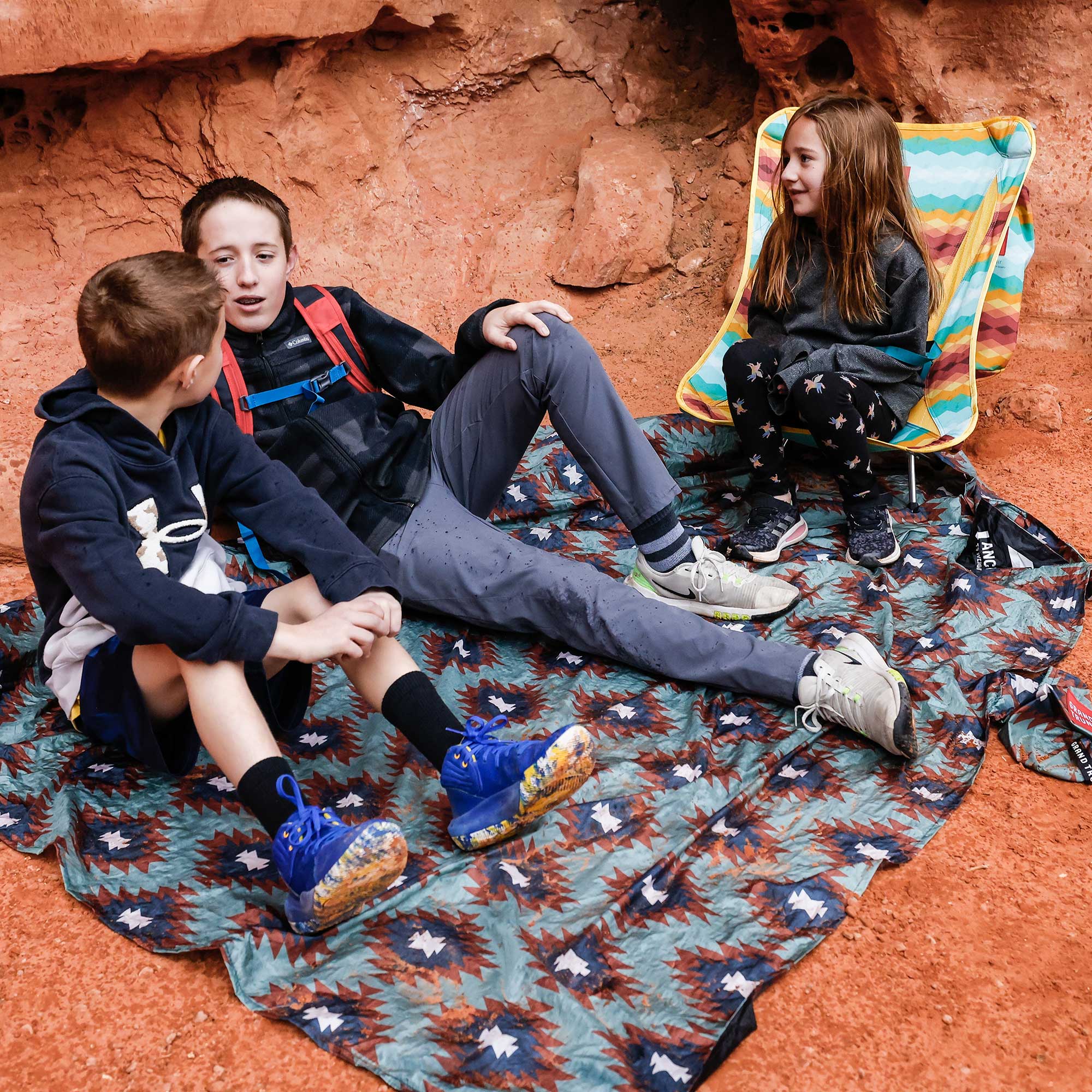 Three children sitting on a patterned blanket in a natural rock setting