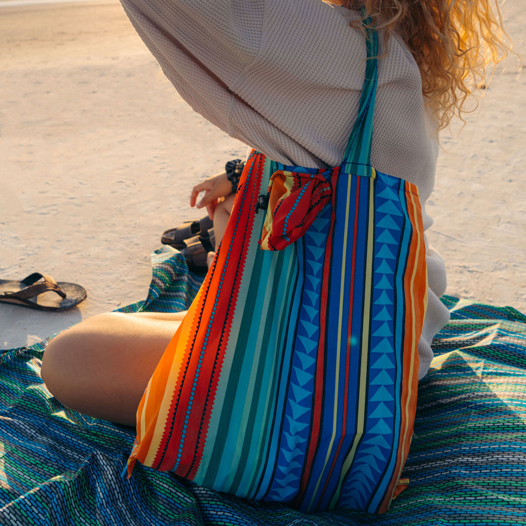 woman sitting on beach holding onto the eco tote beach bag over shoulder carrying beach supplies