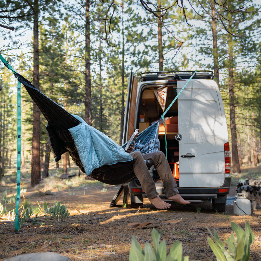 Man reading in evolution hammock suspended from camper van in forest van life propane tank foliage pine barefoot