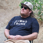 Man wearing a navy 'Grand Trunk' t-shirt with a cap and sunglasses, sitting outdoors.