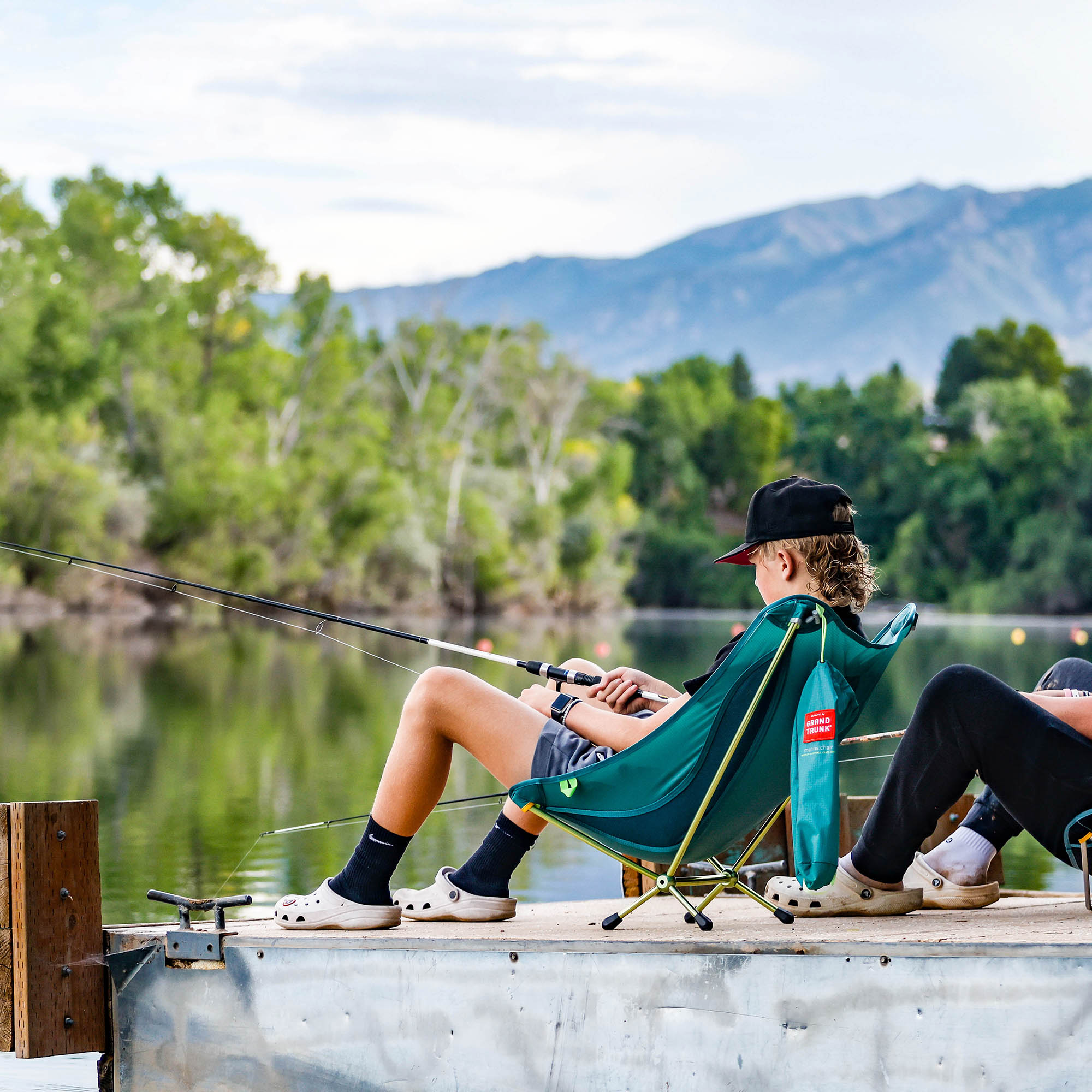 young man sits in mantis chair on dock fishing in a lake with mountains in background