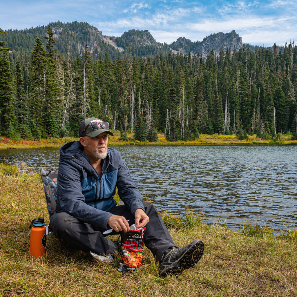 Eating rehydrated camp meal in Grand Trunk Mayfly Chair next to lake and pine trees with mountains in the background