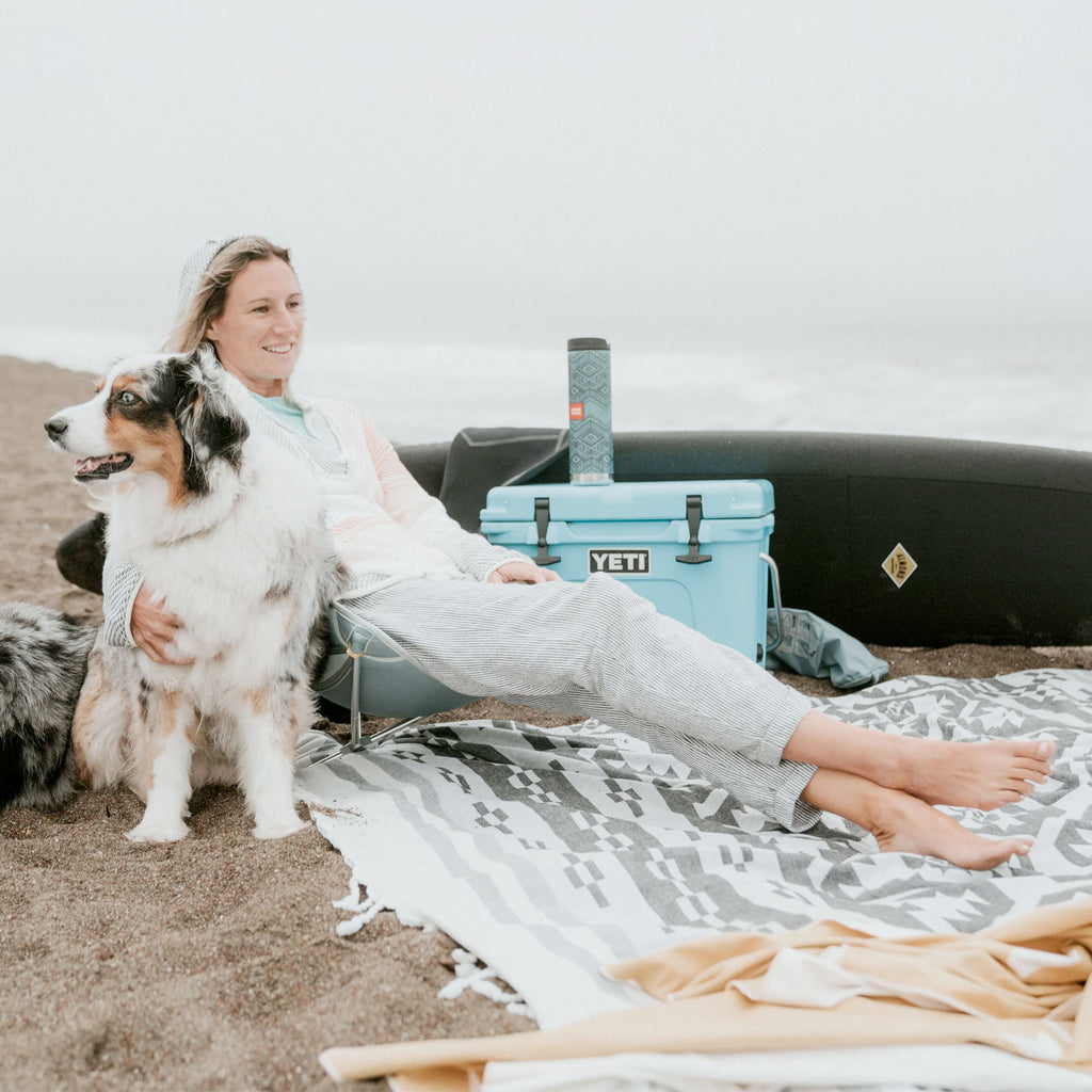 Woman on beach in Grand Trunk Monarch chair relaxing with dog and surfboard with klean kanteen water bottle and yeti cooler