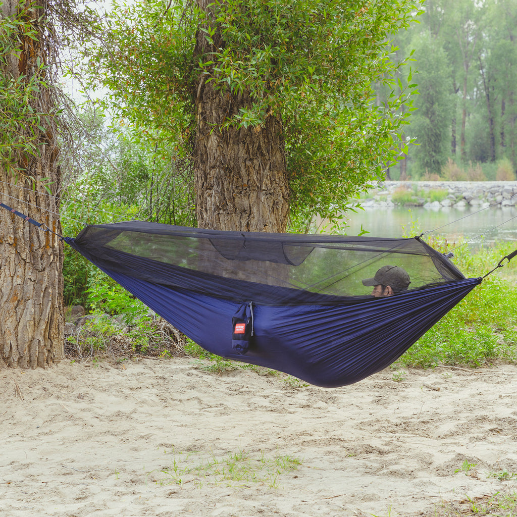 man lays in the skeeter beeter hammock next to a large body of water with big trees and forest