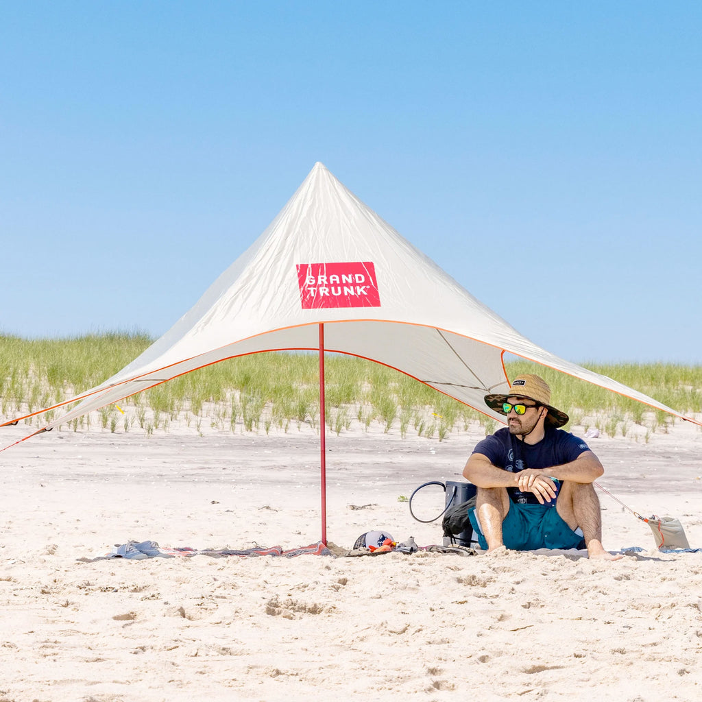 man sits in the shade under the shadecaster 2 on a white sand beach on a clear sky day
