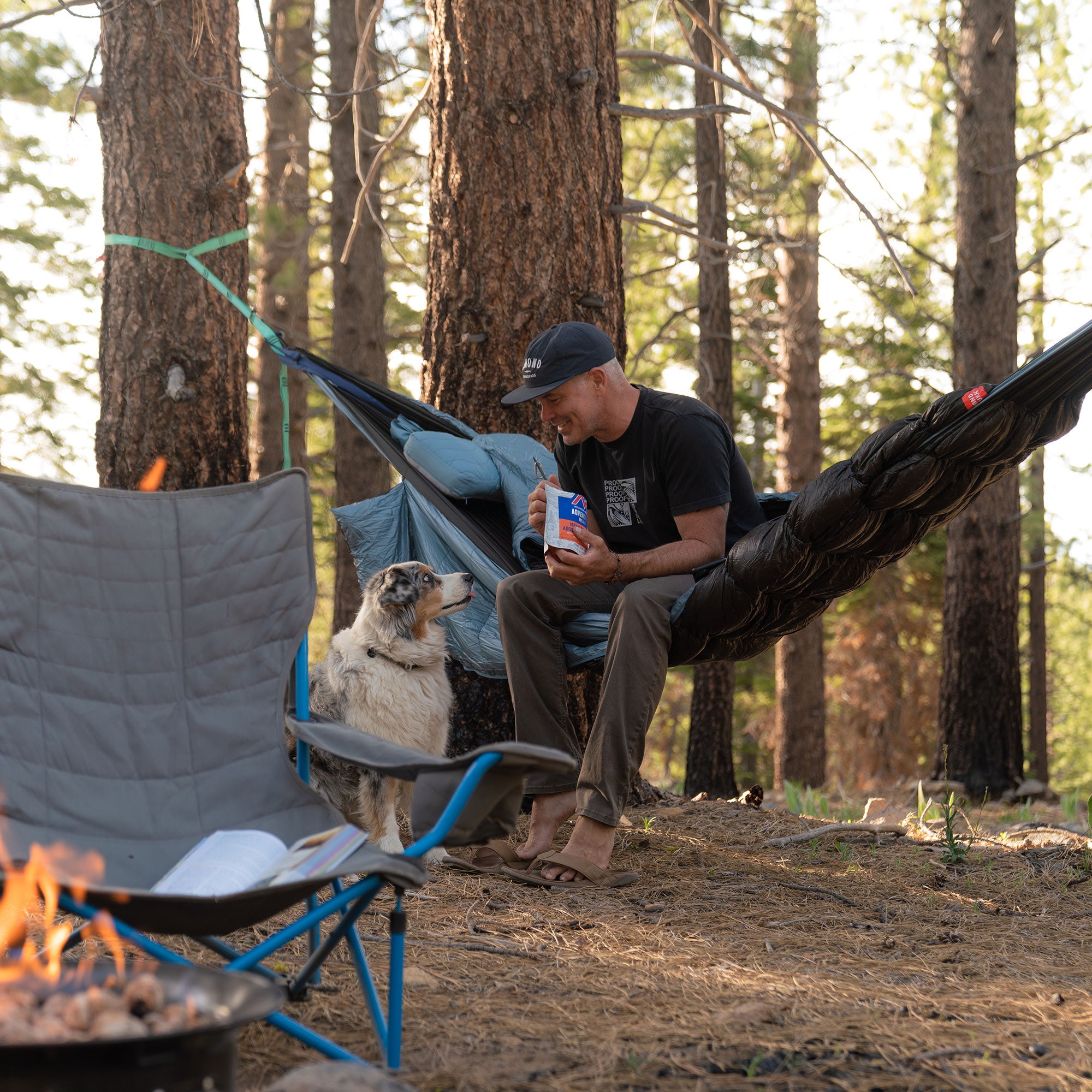 Man lounging in evolution hammock eating mountain house meals by a firepit with his dog looking longingly for some food